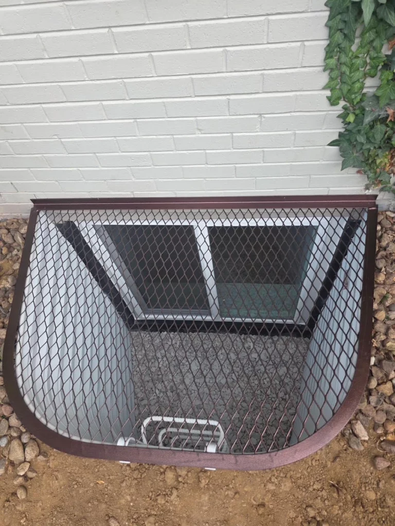 Brown mesh grate over a white window well with ladder and gravel base beside painted brick and ivy in Denver, CO.