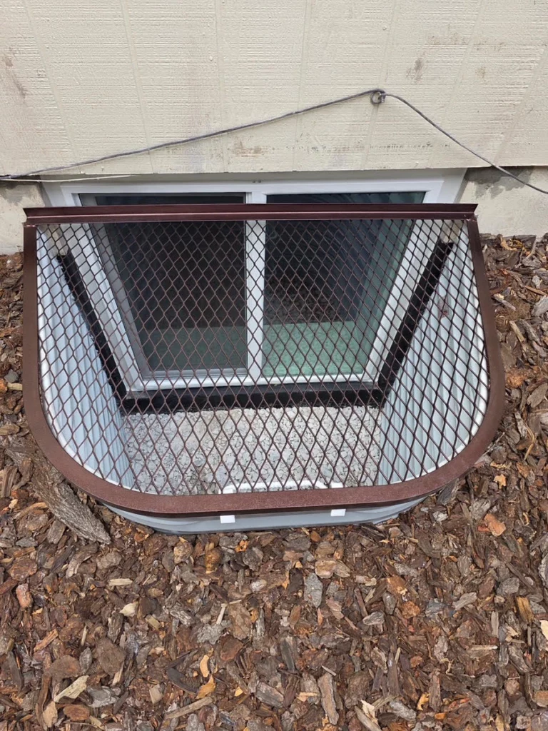 White window well with brown mesh grate and gravel base below a slider window and siding in Lakewood, CO.