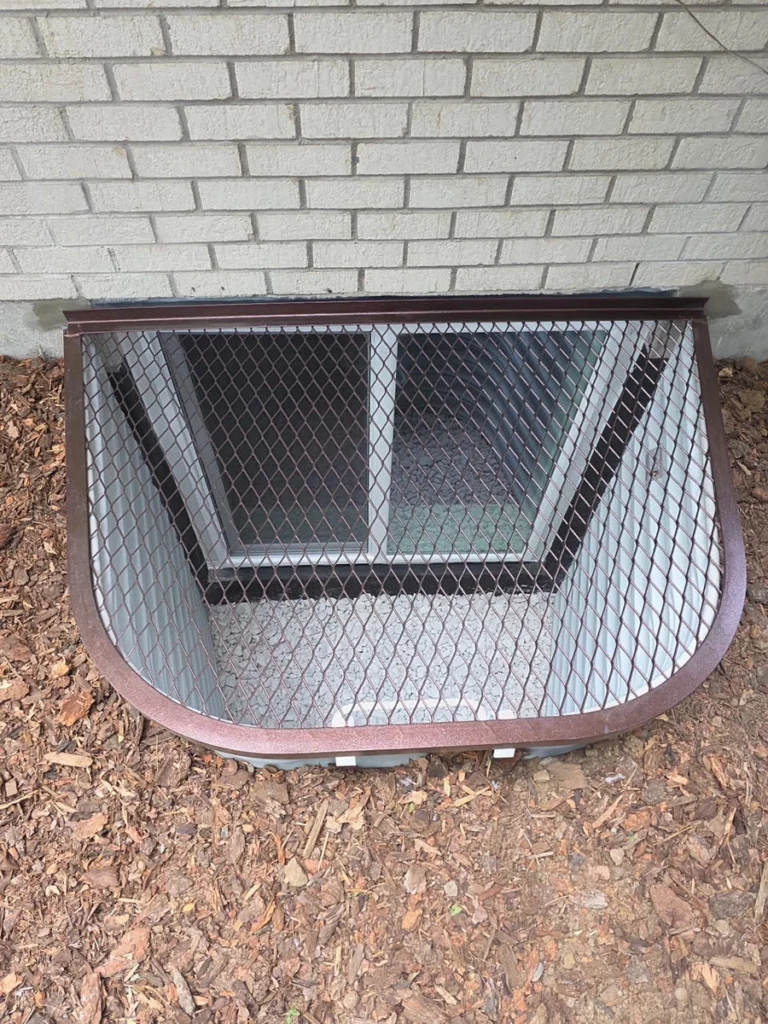 Brown mesh grate over a white window well with gravel base beside painted brick in Lakewood, CO.