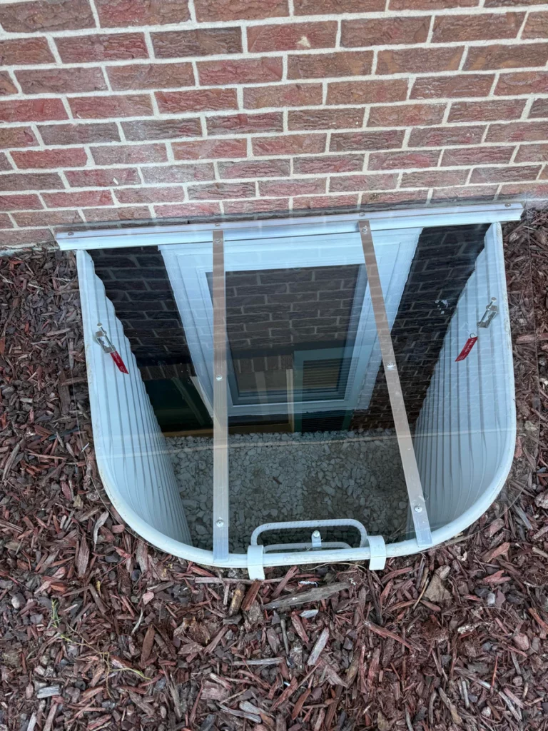 Clear cover over a white window well with ladder and gravel beside red brick in Littleton, CO.